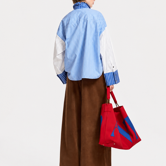 A model is shown from the back wearing a blue and white striped button-down shirt with white sleeves, brown wide-leg trousers, and holding a red tote bag with blue accents, set against a plain white studio background.