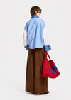 A model is shown from the back wearing a blue and white striped button-down shirt with white sleeves, brown wide-leg trousers, and holding a red tote bag with blue accents, set against a plain white studio background.