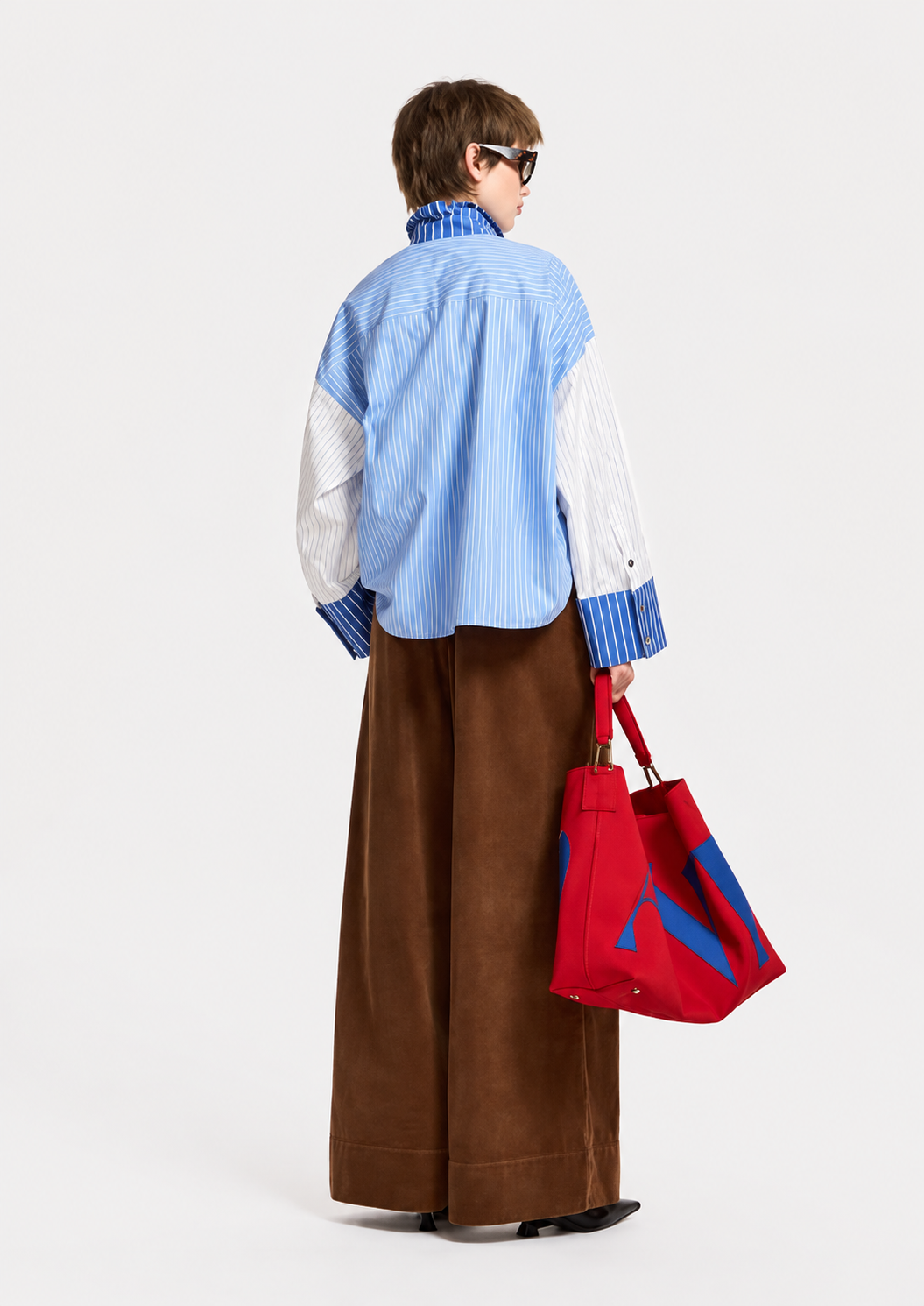 A model is shown from the back wearing a blue and white striped button-down shirt with white sleeves, brown wide-leg trousers, and holding a red tote bag with blue accents, set against a plain white studio background.