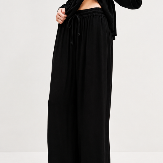 A model wearing black wide-leg drawstring trousers and a black long-sleeve top, shown from the chest down against a plain light gray studio background.