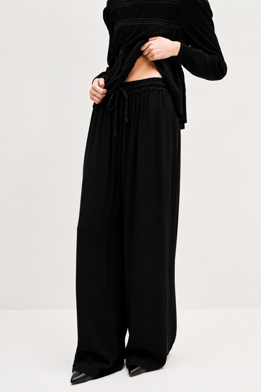A model wearing black wide-leg drawstring trousers and a black long-sleeve top, shown from the chest down against a plain light gray studio background.