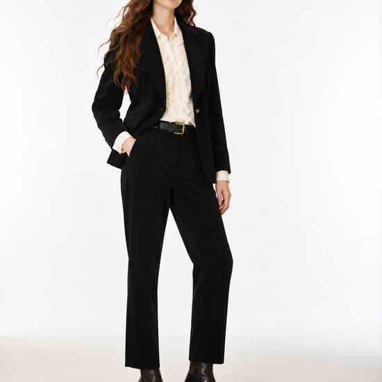 A woman modeling a black tailored suit consisting of a blazer and trousers, worn over a cream patterned blouse with a black belt and black ankle boots, photographed against a plain light gray studio background.