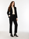 A woman modeling a black tailored suit consisting of a blazer and trousers, worn over a cream patterned blouse with a black belt and black ankle boots, photographed against a plain light gray studio background.