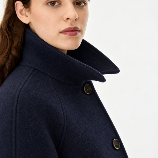 Close-up of a woman wearing a dark navy wool coat with a high structured collar and dark buttons, photographed against a clean white studio background.