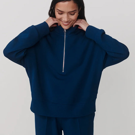 A woman wearing a navy blue half-zip sweatshirt and matching navy blue sweatpants, posing against a light gray studio background.