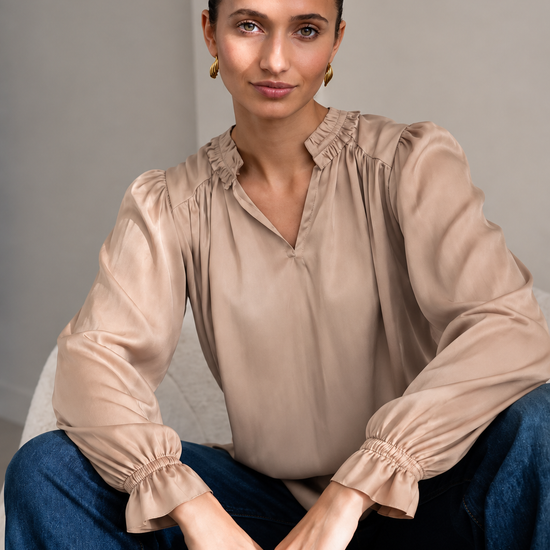 A woman wearing a beige long-sleeve blouse with gathered ruffles at the collar and cuffs, paired with blue jeans, sitting on a light gray upholstered chair.