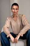 A woman wearing a beige long-sleeve blouse with gathered ruffles at the collar and cuffs, paired with blue jeans, sitting on a light gray upholstered chair.