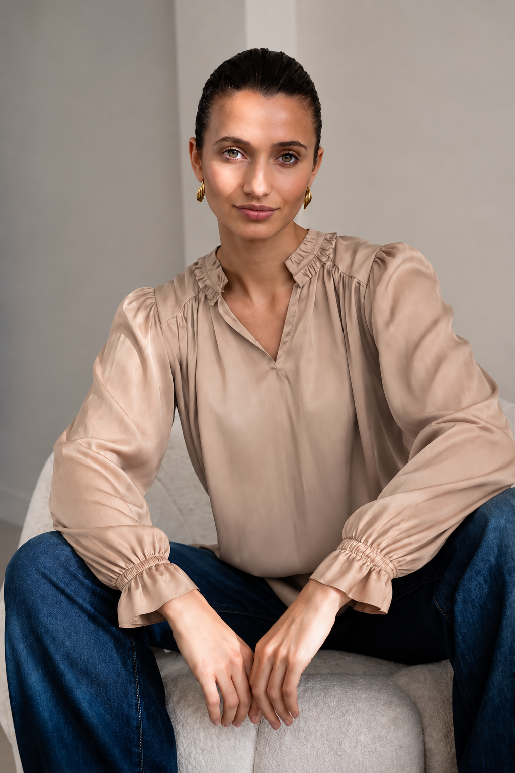 A woman wearing a beige long-sleeve blouse with gathered ruffles at the collar and cuffs, paired with blue jeans, sitting on a light gray upholstered chair.