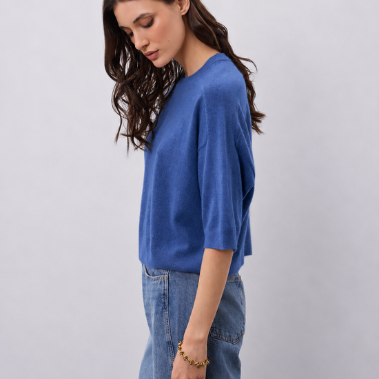 A woman wearing a blue knit top with three-quarter sleeves and light blue jeans, standing against a plain light gray studio background.