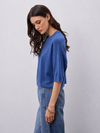 A woman wearing a blue knit top with three-quarter sleeves and light blue jeans, standing against a plain light gray studio background.