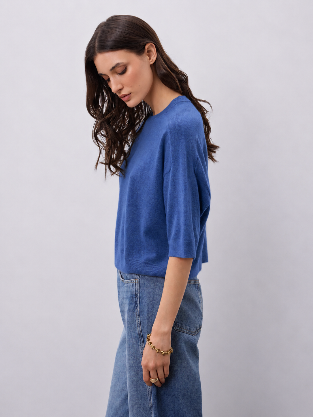 A woman wearing a blue knit top with three-quarter sleeves and light blue jeans, standing against a plain light gray studio background.