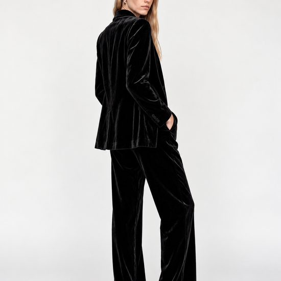 A woman modeling a black velvet two-piece suit consisting of a cropped blazer and wide-leg trousers, paired with black pointed-toe heels, against a plain white studio background.