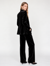 A woman modeling a black velvet two-piece suit consisting of a cropped blazer and wide-leg trousers, paired with black pointed-toe heels, against a plain white studio background.