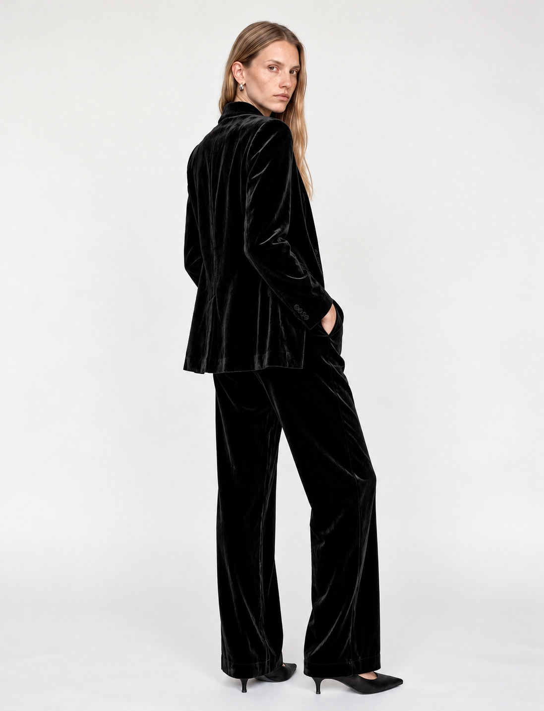 A woman modeling a black velvet two-piece suit consisting of a cropped blazer and wide-leg trousers, paired with black pointed-toe heels, against a plain white studio background.