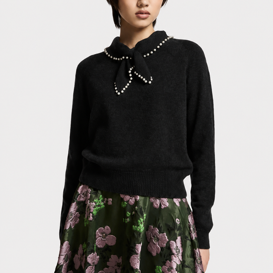 A model wearing a black knit sweater with a pearl-trimmed bow collar and a dark green floral skirt, photographed against a light gray studio background.
