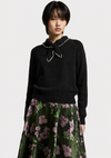 A model wearing a black knit sweater with a pearl-trimmed bow collar and a dark green floral skirt, photographed against a light gray studio background.
