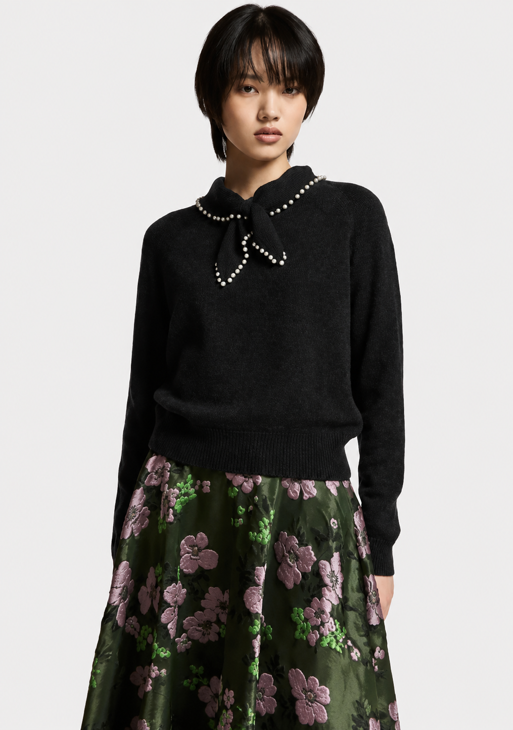A model wearing a black knit sweater with a pearl-trimmed bow collar and a dark green floral skirt, photographed against a light gray studio background.