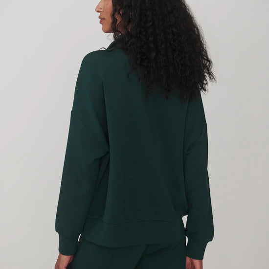 A woman with long, curly black hair is shown from the back wearing a dark forest green sweatshirt and matching sweatpants against a plain light gray studio background.