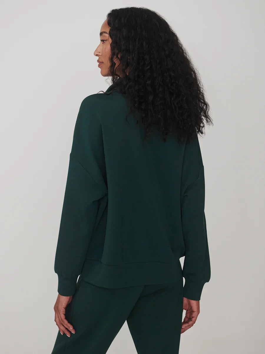A woman with long, curly black hair is shown from the back wearing a dark forest green sweatshirt and matching sweatpants against a plain light gray studio background.