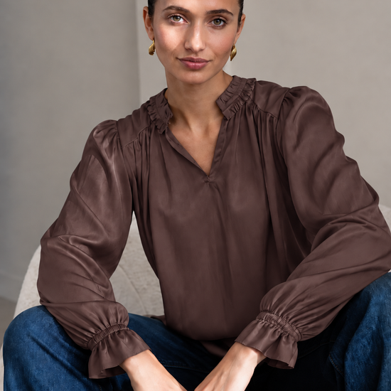 A woman wearing a brown long-sleeve blouse with gathered ruffles at the collar and cuffs, paired with blue jeans, sitting on a light gray upholstered chair.