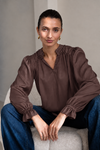 A woman wearing a brown long-sleeve blouse with gathered ruffles at the collar and cuffs, paired with blue jeans, sitting on a light gray upholstered chair.