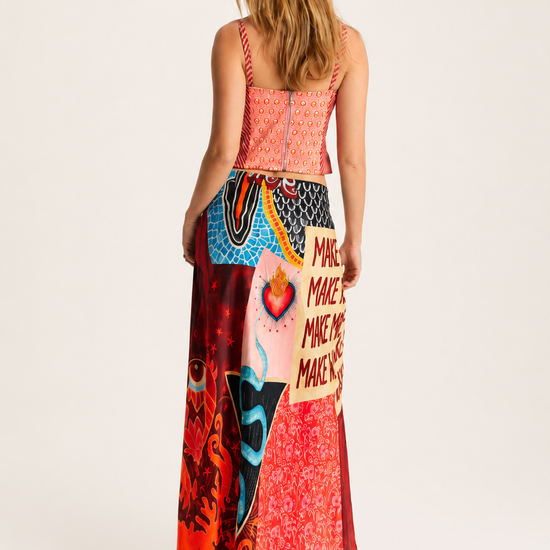 A woman is shown from the back wearing a long, colorful patchwork maxi dress with a coral pink bodice and a vibrant multicolor skirt featuring various graphic and patterned panels.