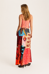 A woman is shown from the back wearing a long, colorful patchwork maxi dress with a coral pink bodice and a vibrant multicolor skirt featuring various graphic and patterned panels.