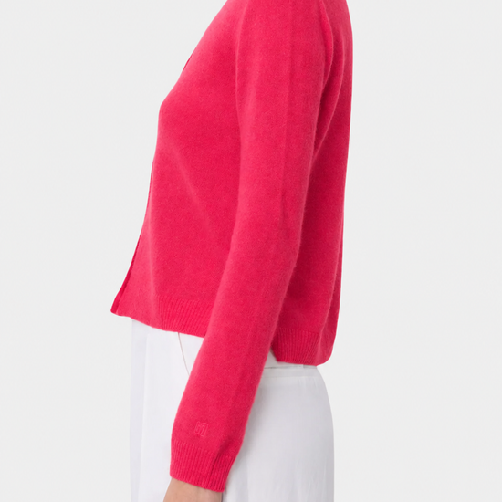 A woman shown in side profile wearing a bright pink cropped knit cardigan with long sleeves and white pants against a light gray studio background.