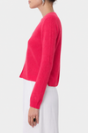 A woman shown in side profile wearing a bright pink cropped knit cardigan with long sleeves and white pants against a light gray studio background.