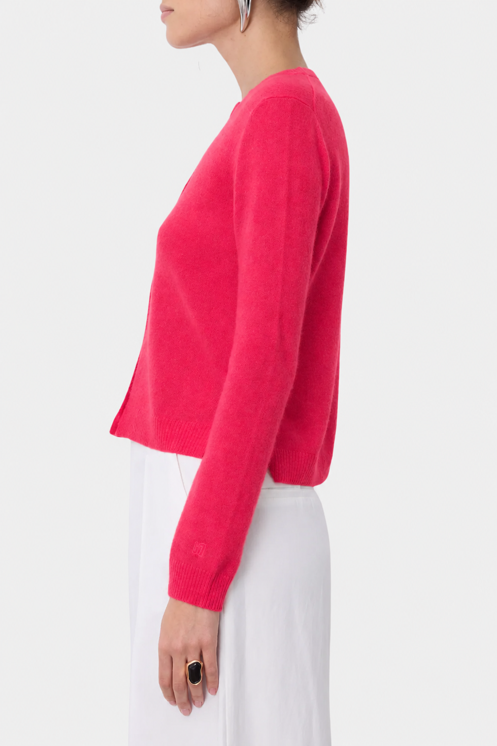 A woman shown in side profile wearing a bright pink cropped knit cardigan with long sleeves and white pants against a light gray studio background.