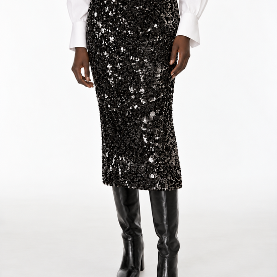 A black sequined midi skirt worn by a model, styled with a white long-sleeve blouse and black knee-high boots against a plain white studio background.