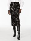 A black sequined midi skirt worn by a model, styled with a white long-sleeve blouse and black knee-high boots against a plain white studio background.