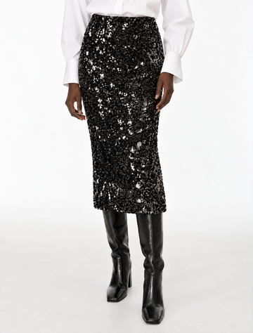 A black sequined midi skirt worn by a model, styled with a white long-sleeve blouse and black knee-high boots against a plain white studio background.