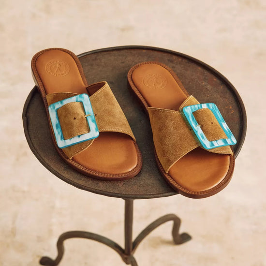 Pair of brown sandals with turquoise buckles on a metal stand.