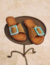 Pair of brown sandals with turquoise buckles on a metal stand.
