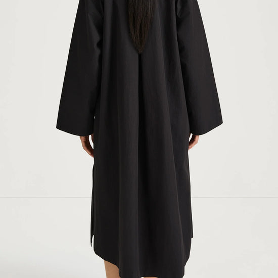 A person shown from the back wearing a loose, long black coat or dress over a white top, paired with black shoes, against a plain light studio background.