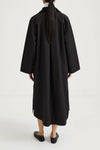 A person shown from the back wearing a loose, long black coat or dress over a white top, paired with black shoes, against a plain light studio background.