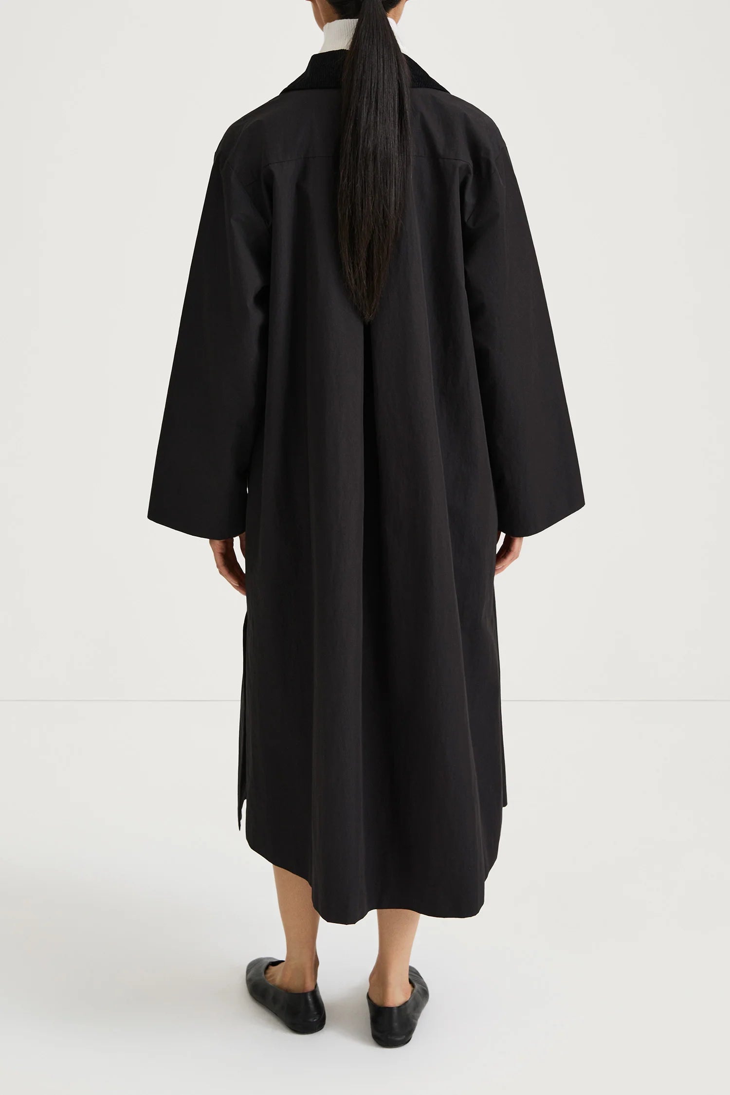 A person shown from the back wearing a loose, long black coat or dress over a white top, paired with black shoes, against a plain light studio background.