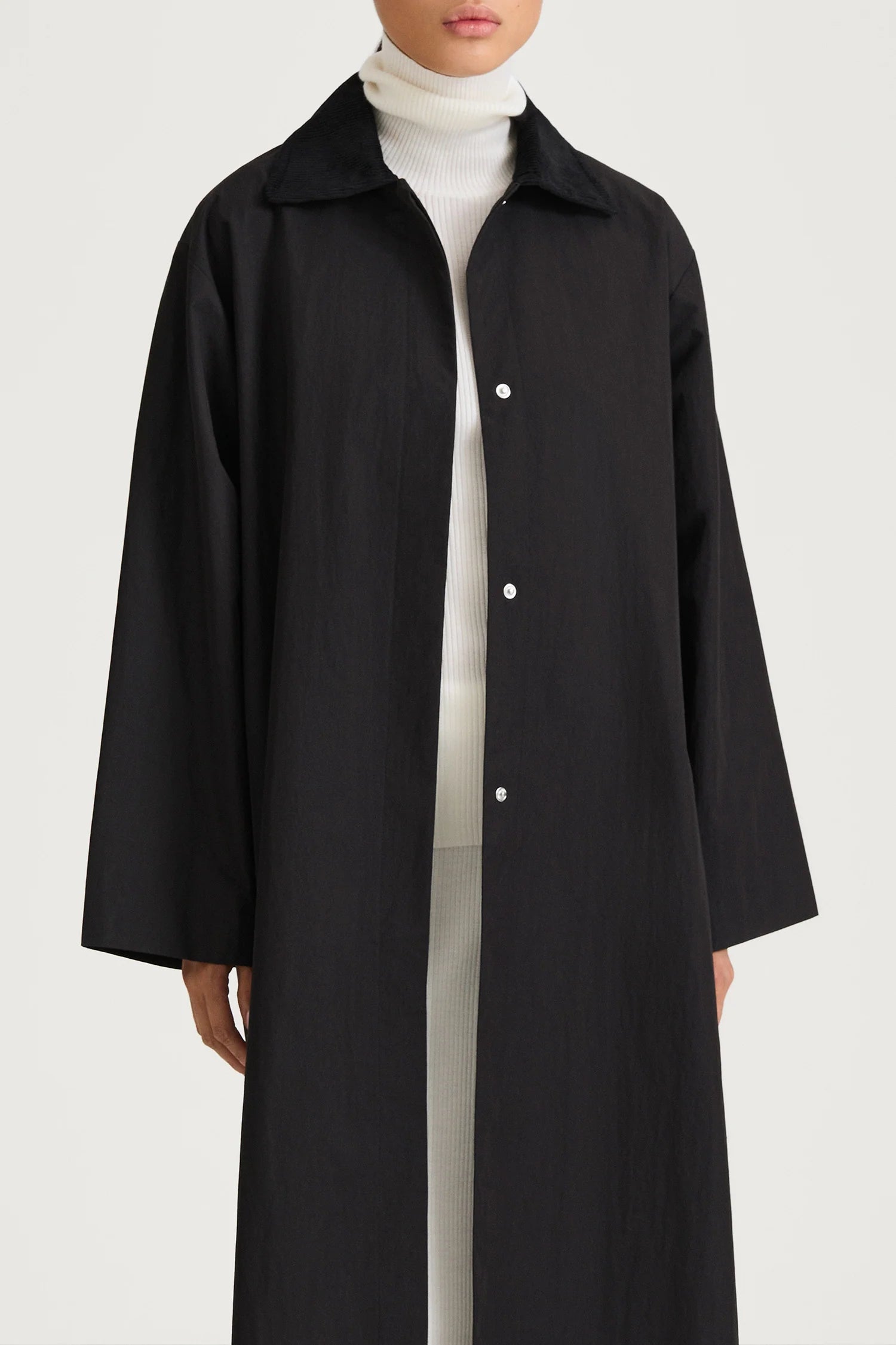 A model wearing a long black button-up coat over a white turtleneck sweater, shown from the neck down against a plain light gray studio background.