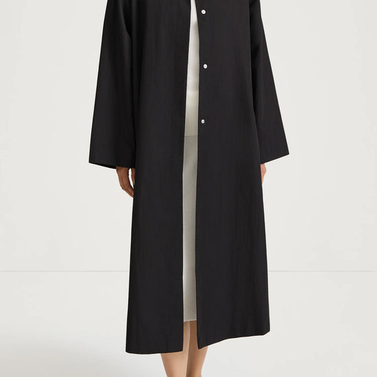 A model wearing a long black button-front coat over a white turtleneck and light dress, paired with black flats, shot against a plain white studio background.