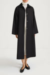 A model wearing a long black button-front coat over a white turtleneck and light dress, paired with black flats, shot against a plain white studio background.