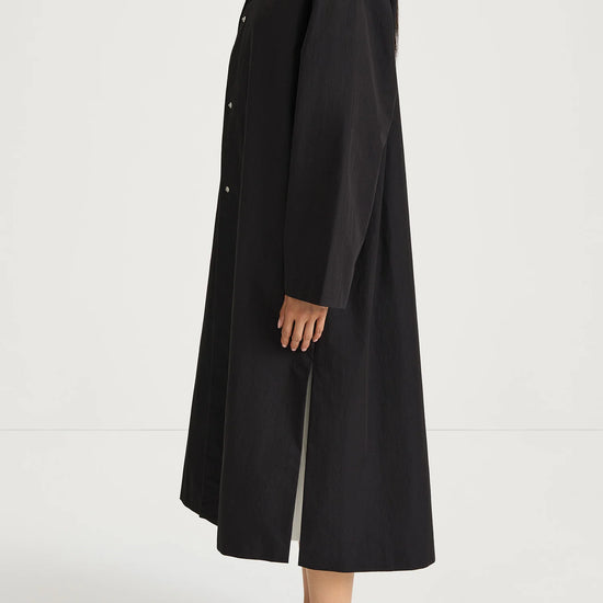 A model shown from the side wearing a long black coat over a white mock-neck top and black flats, set against a plain white studio background.