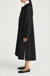 A model shown from the side wearing a long black coat over a white mock-neck top and black flats, set against a plain white studio background.
