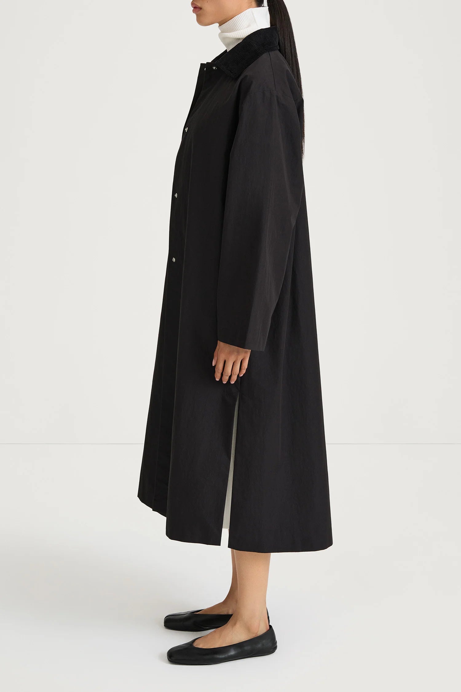 A model shown from the side wearing a long black coat over a white mock-neck top and black flats, set against a plain white studio background.