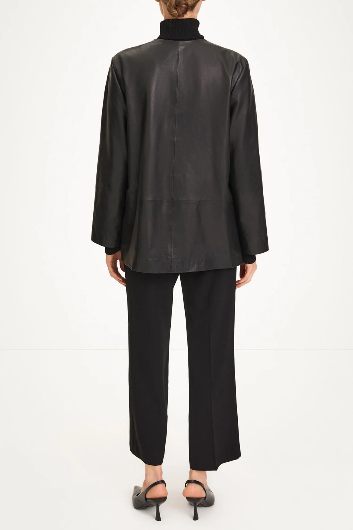 A model shown from the back wearing a black leather jacket with a high mock neck, black wide-leg trousers, and black high-heeled sandals against a plain white studio background.