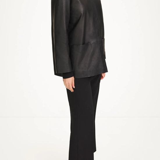A model wearing a black leather long-sleeve turtleneck jacket, black trousers, and black pointed-toe heels, shown from a side profile against a plain white studio background.