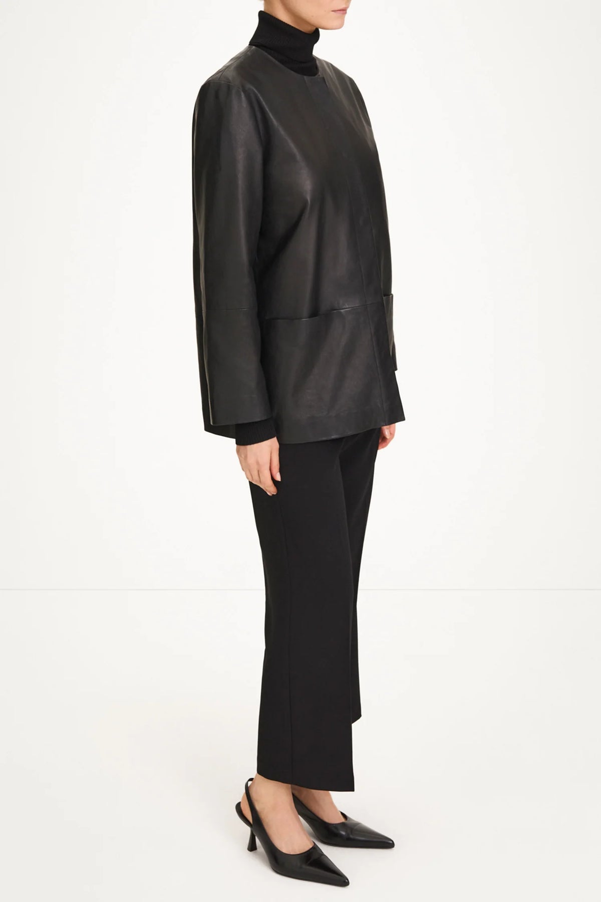 A model wearing a black leather long-sleeve turtleneck jacket, black trousers, and black pointed-toe heels, shown from a side profile against a plain white studio background.