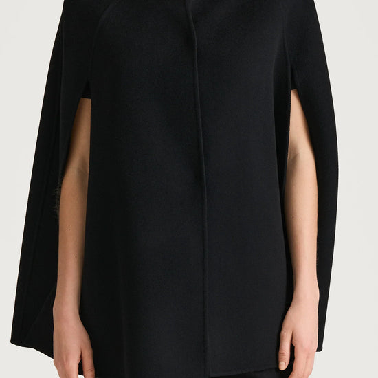 A model wearing a black cape-style wrap coat with a mock neck and short sleeves, paired with black trousers, against a light gray studio background.