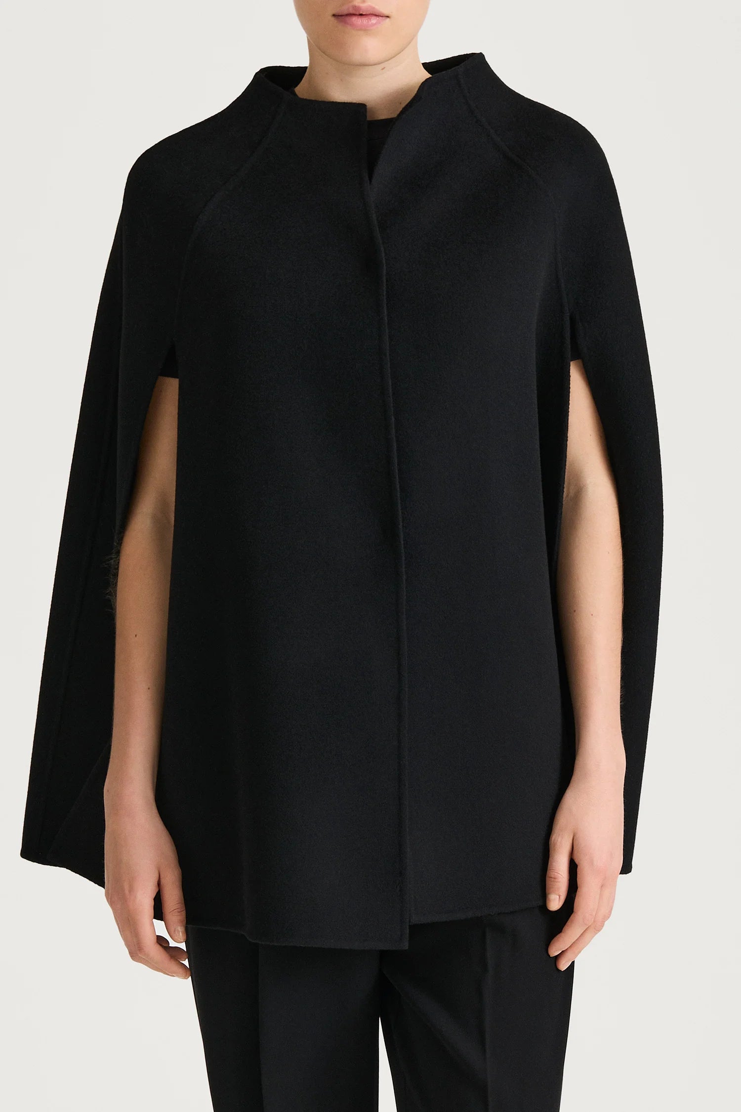 A model wearing a black cape-style wrap coat with a mock neck and short sleeves, paired with black trousers, against a light gray studio background.