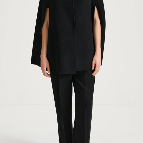 A model wearing a black sleeveless cape-style top with side slits and matching black wide-leg trousers, standing against a plain light gray studio background.